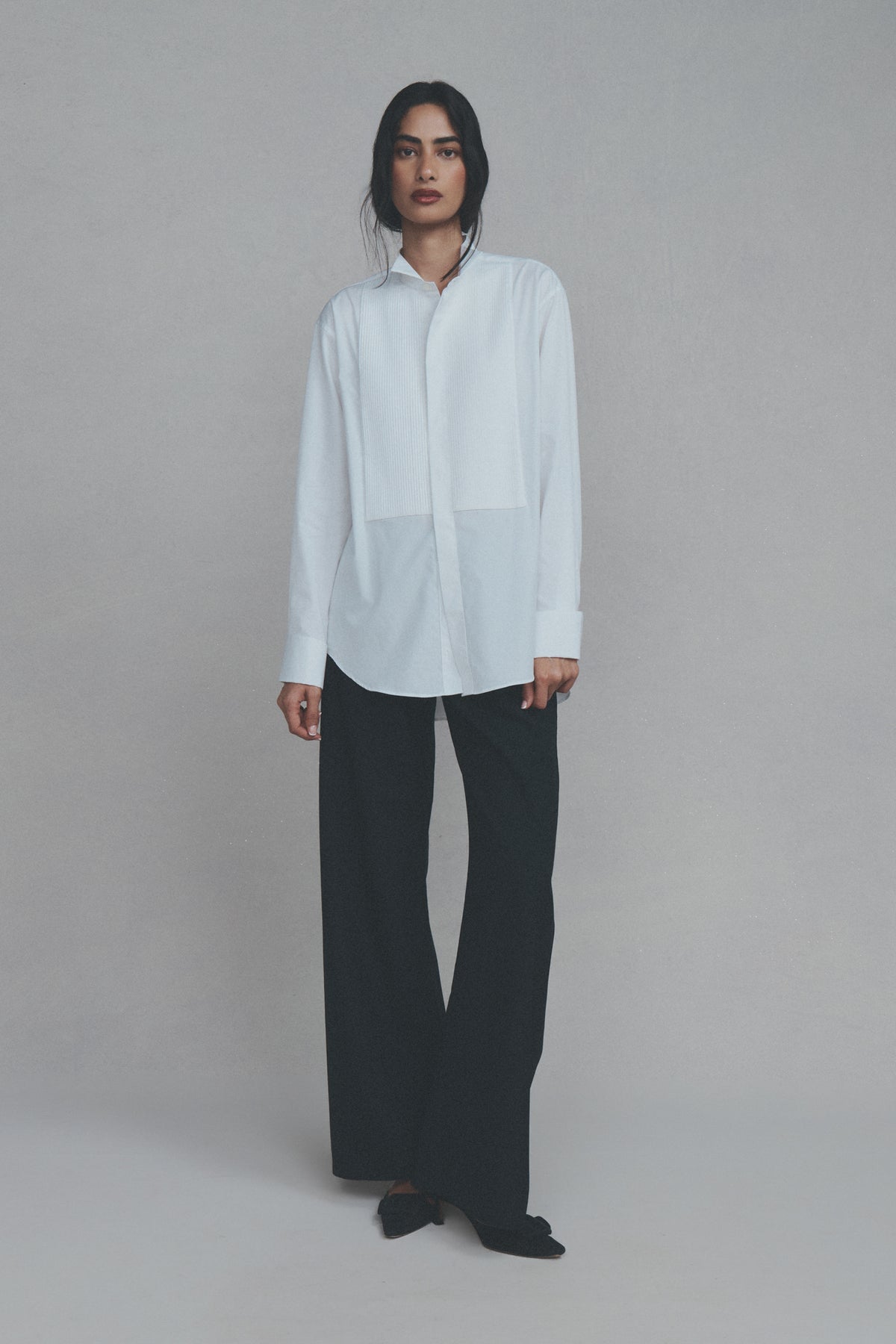 A model stands against a neutral gray studio background wearing a white Comme Si long-sleeve button-up shirt with a unique asymmetrical front panel design and midnight navy bias-cut pants.