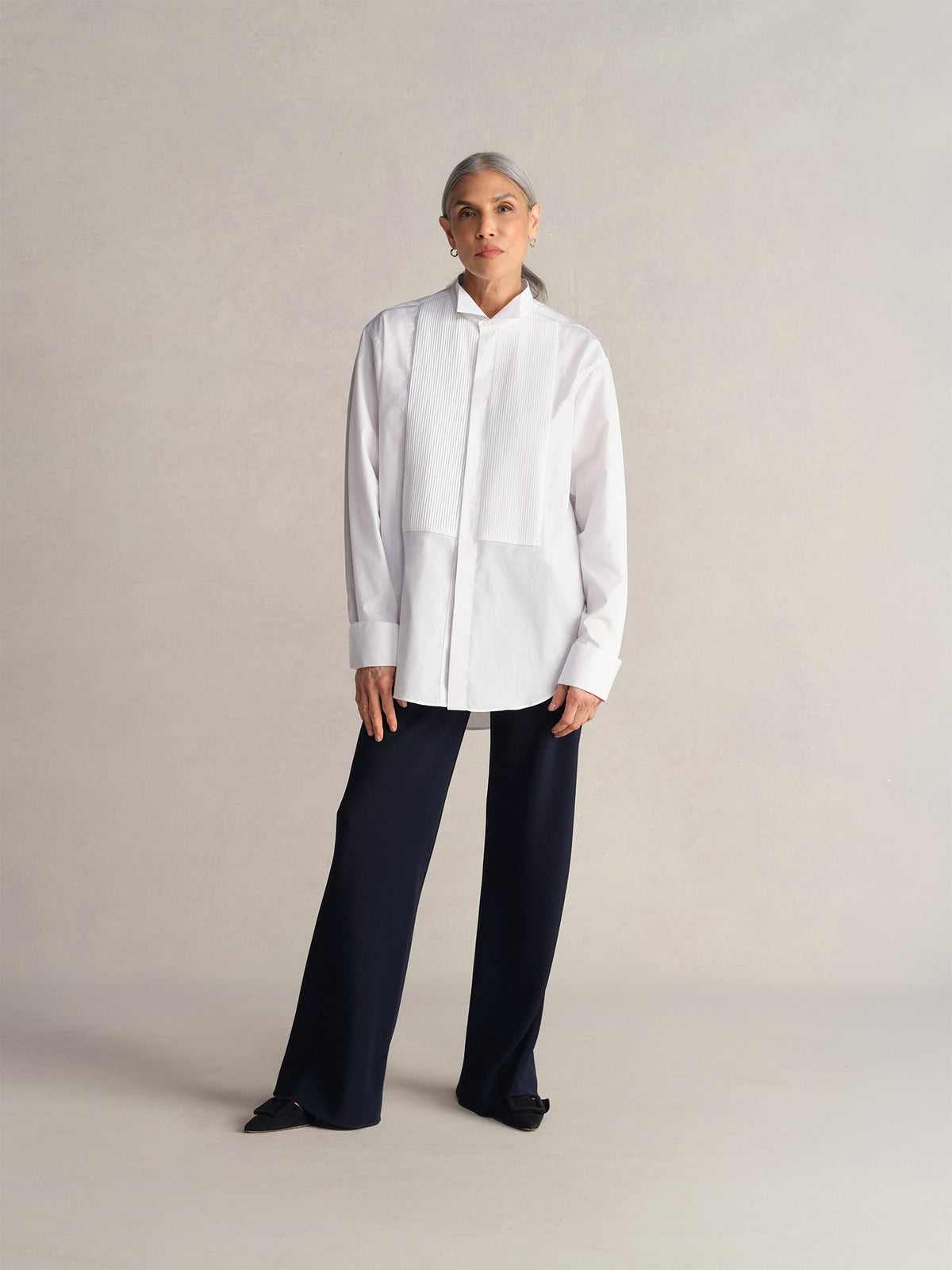 A model stands against a neutral beige studio backdrop wearing a Comme Si white long-sleeve button-up shirt with a high collar and midnight bias-cut pants.