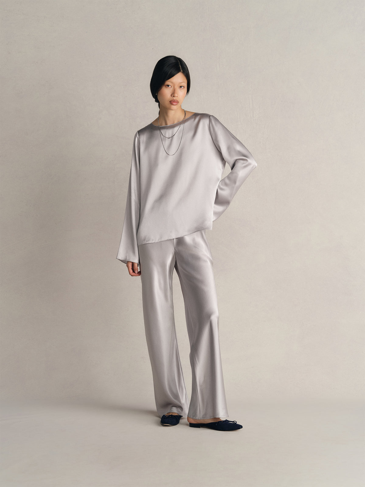Model wearing a Comme Si silk silver bell-sleeve top and matching silk bias pants with layered necklaces and black flats.