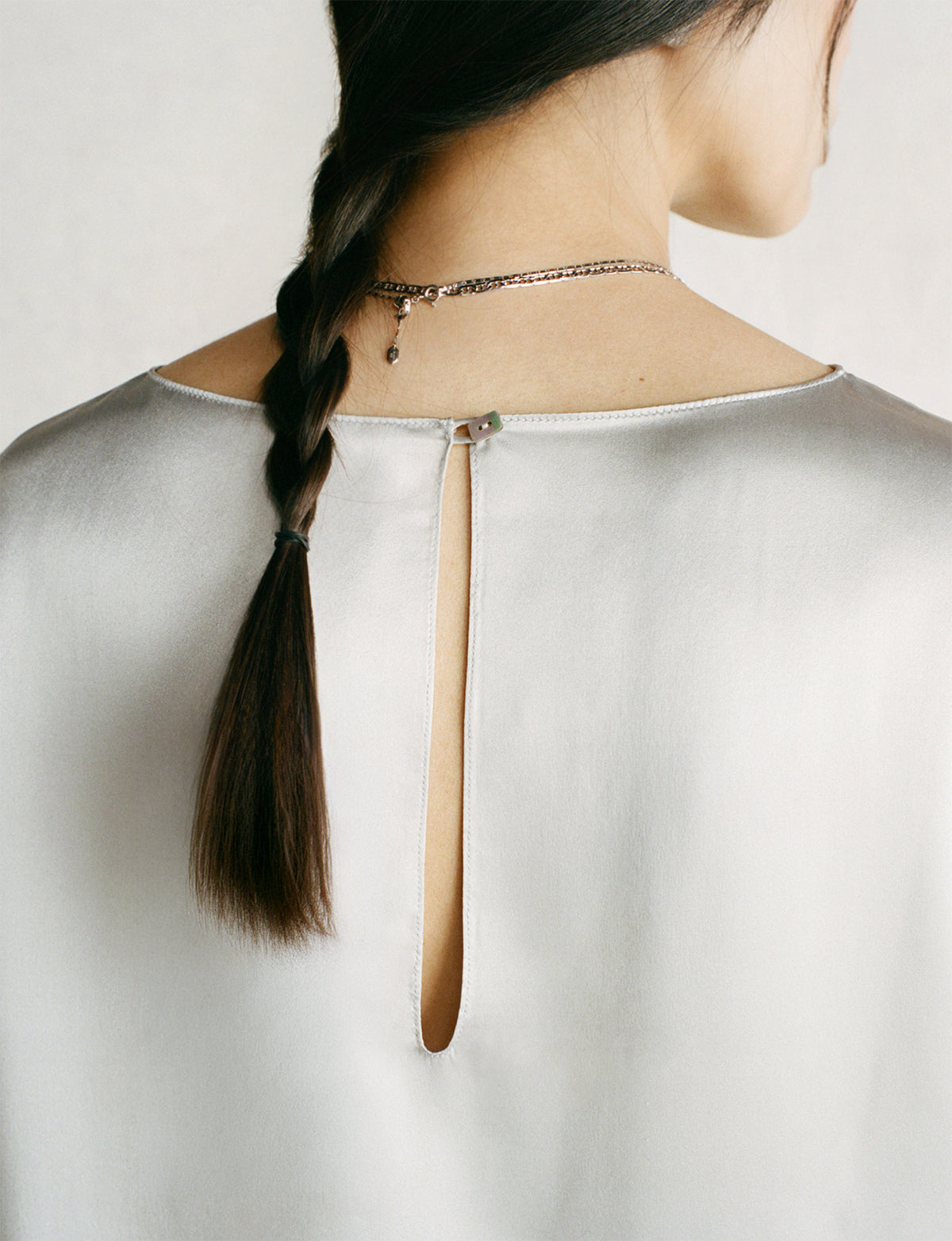 Close-up back view of a woman wearing a Comme Si silver silk top with a keyhole back cutout, styled with a braided ponytail and a delicate silver necklace.