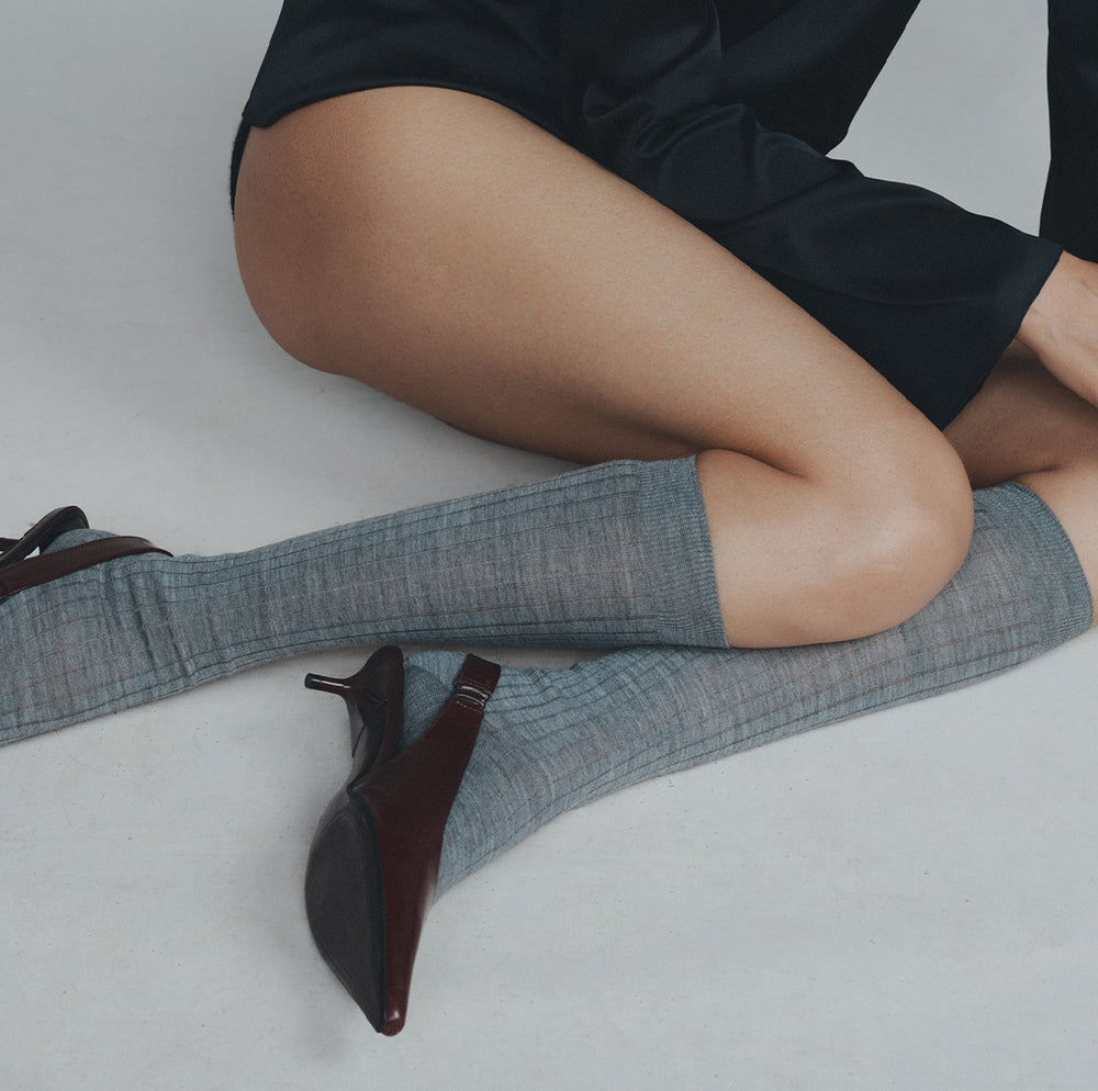 Seated model wearing Comme Si gray ribbed knee-high socks with dark burgundy slingback heels.