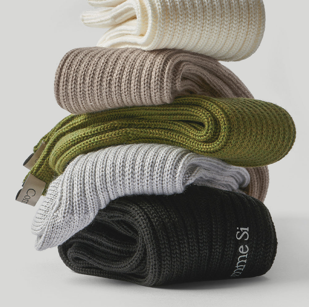 Stack of Comme Si merino wool chunky ribbed socks in cream, beige, olive green, light gray, and black.