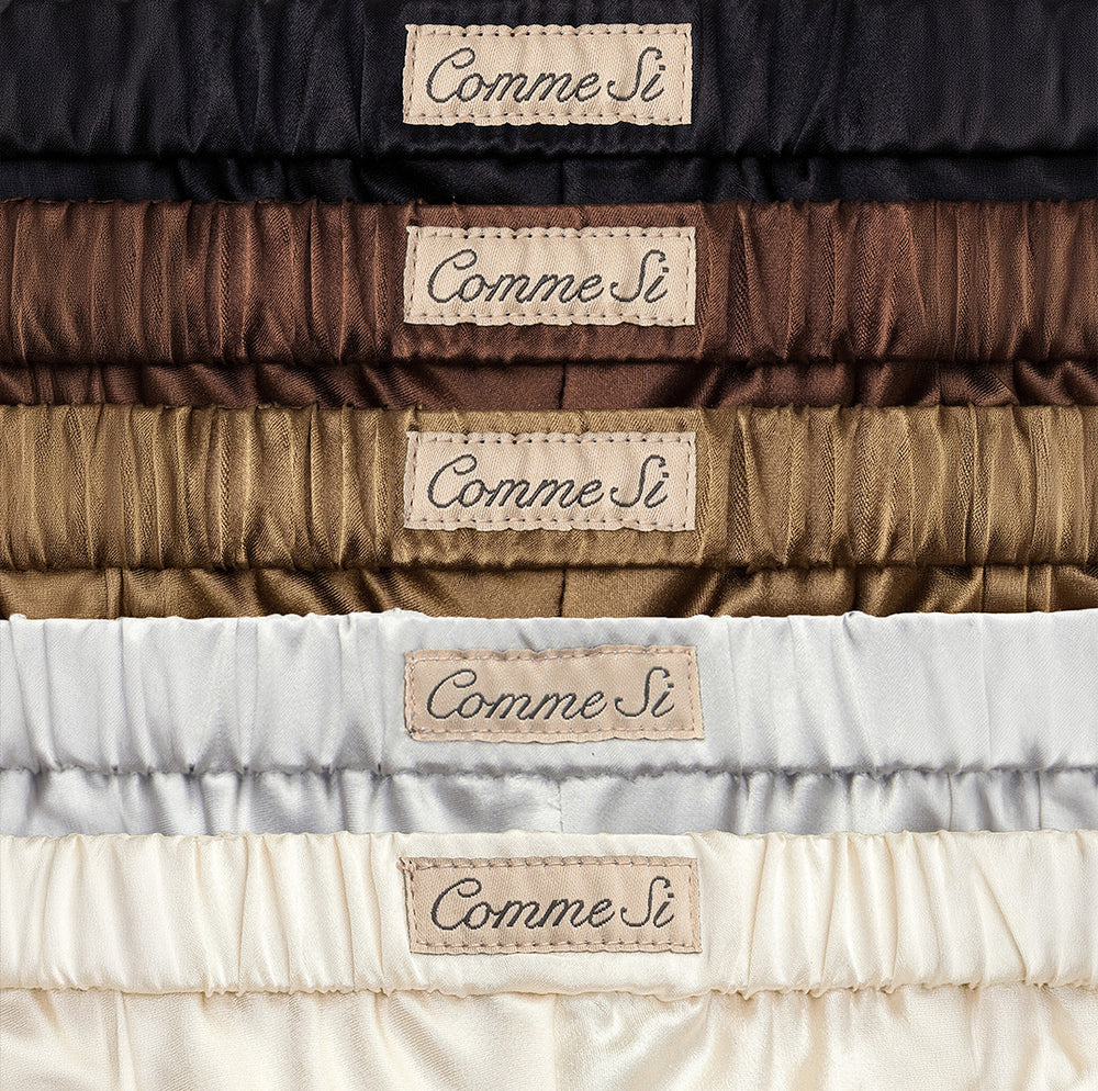 Stacked silk pants in black, espresso brown, taupe, silver, and ivory, showing elastic waistbands and Comme Si labels.