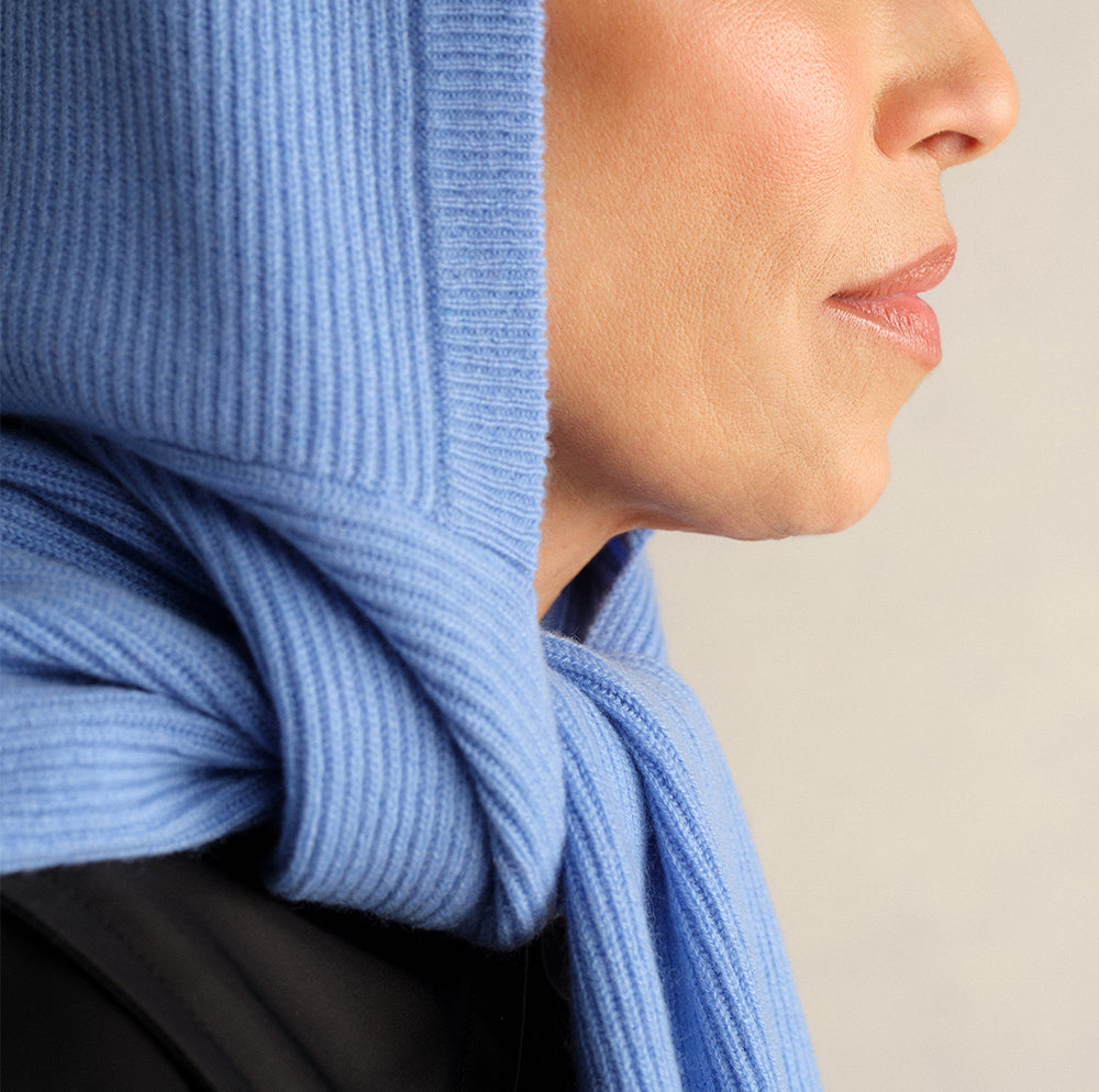 Close-up of a model wearing a Comme Si capri blue ribbed knit scarf wrapped over the head and tied at the neck.