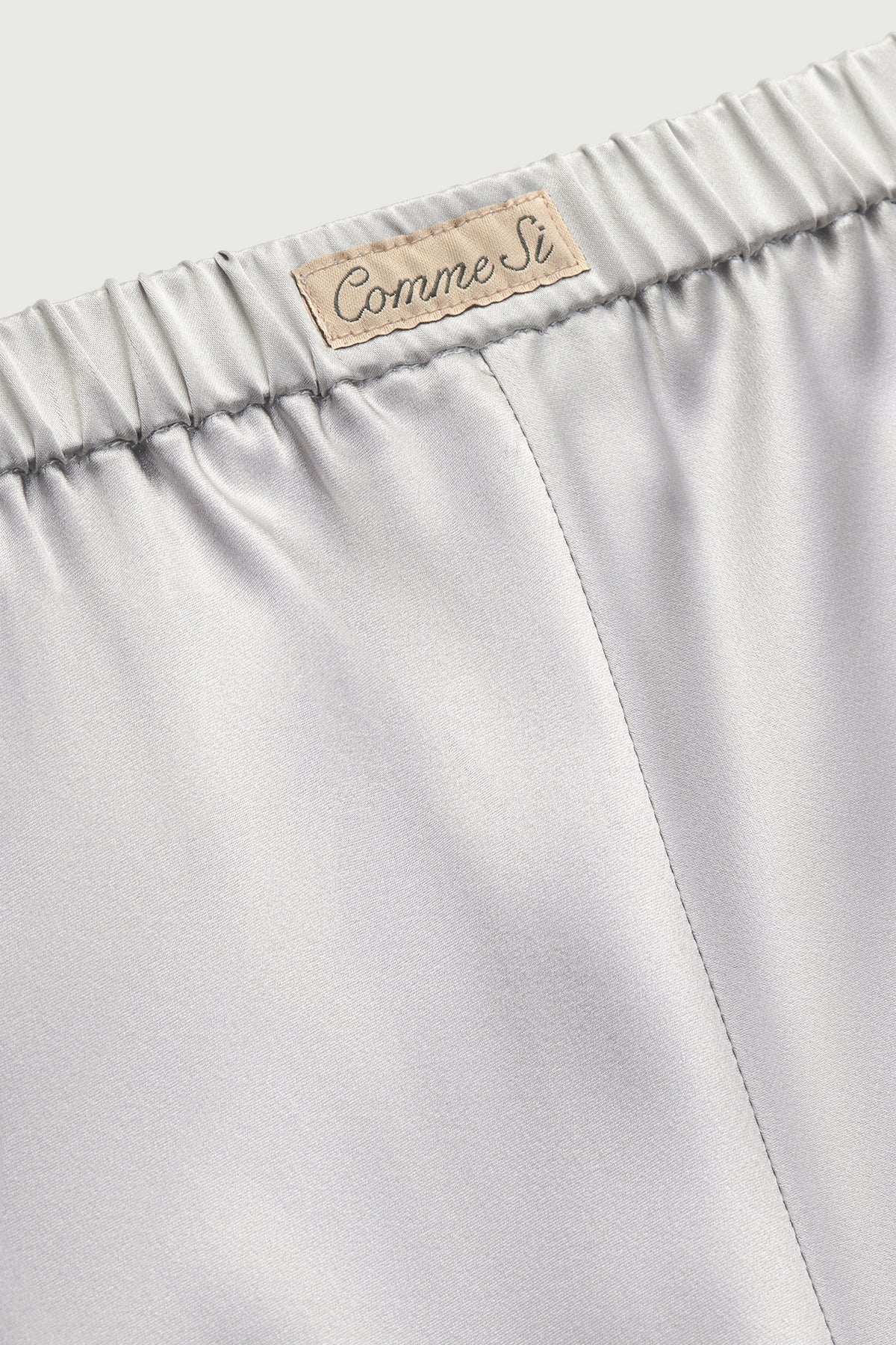 Close-up of silver silk bias-cut pants showing the gathered elastic waistband, Comme Si label, and center seam.