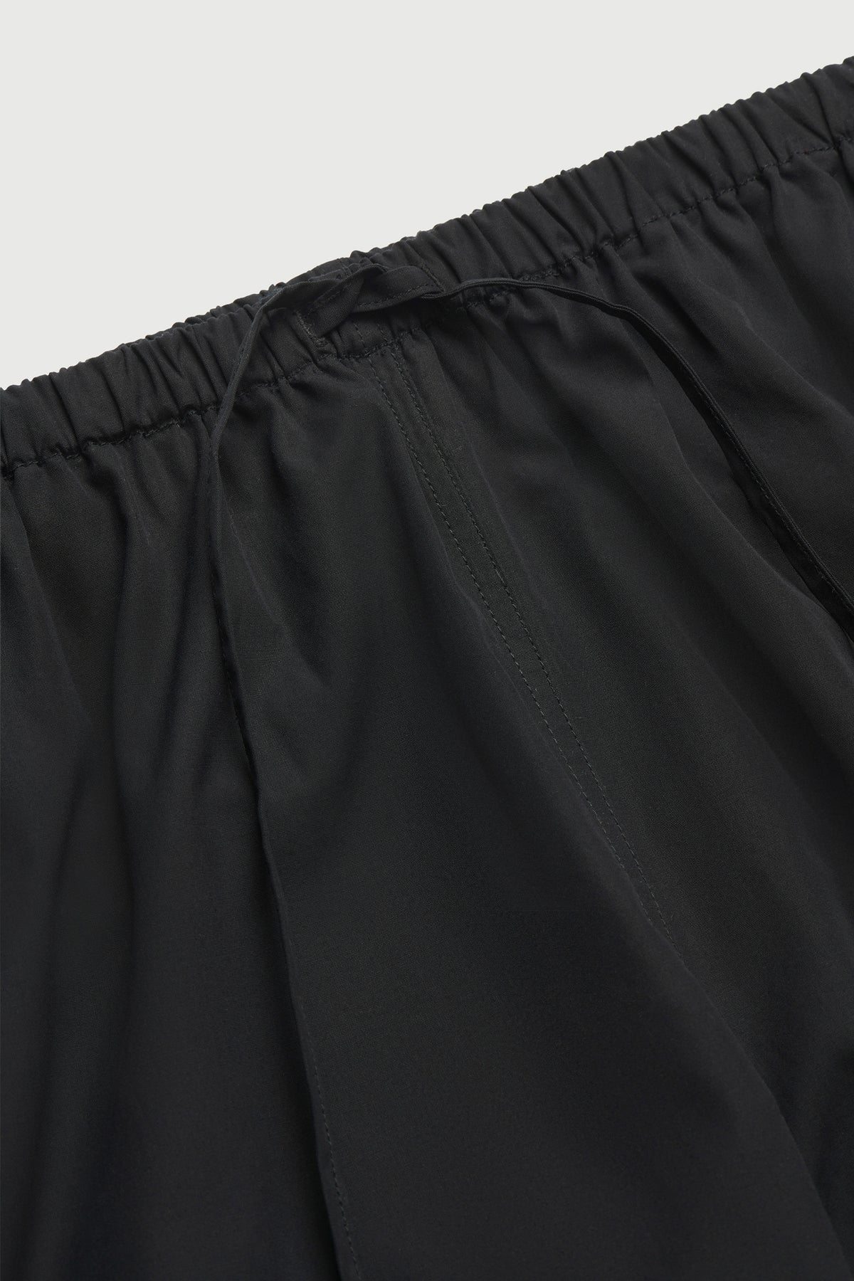 Close-up detail of Comme Si black cotton poplin pants showing the elasticized drawstring waistband and fabric texture.