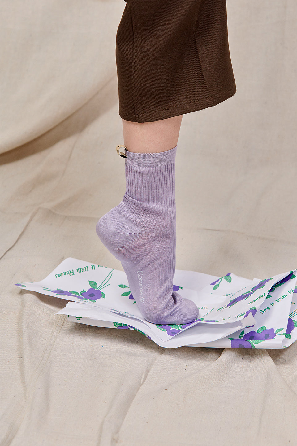 The Agnelli sock in Lilac light purple, foot stepping on a piece of floral bouquet wrapping paper, wearing brown trousers